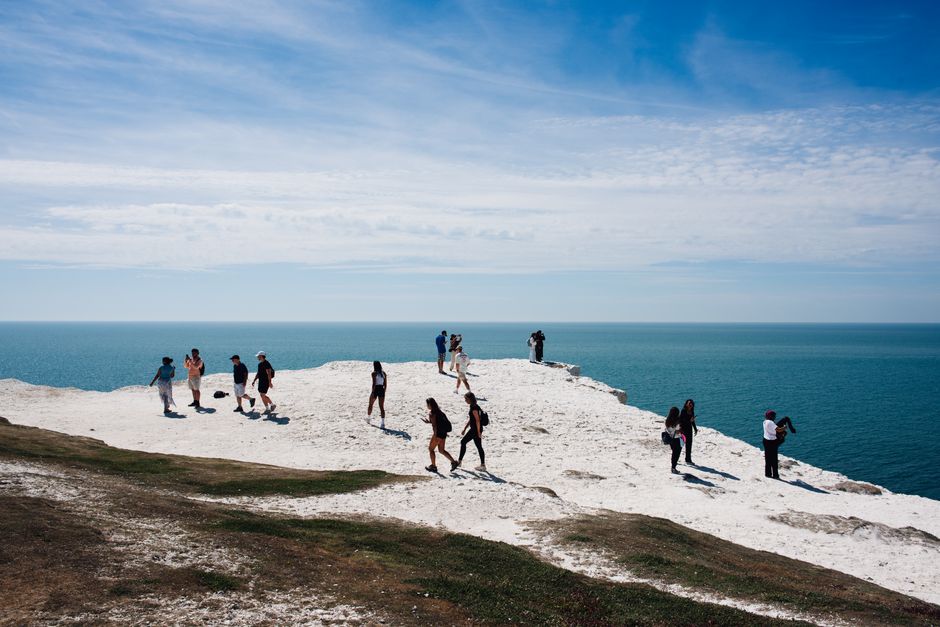A group of people walk along dramatic white cliffs overlooking the ocean under a blue sky.