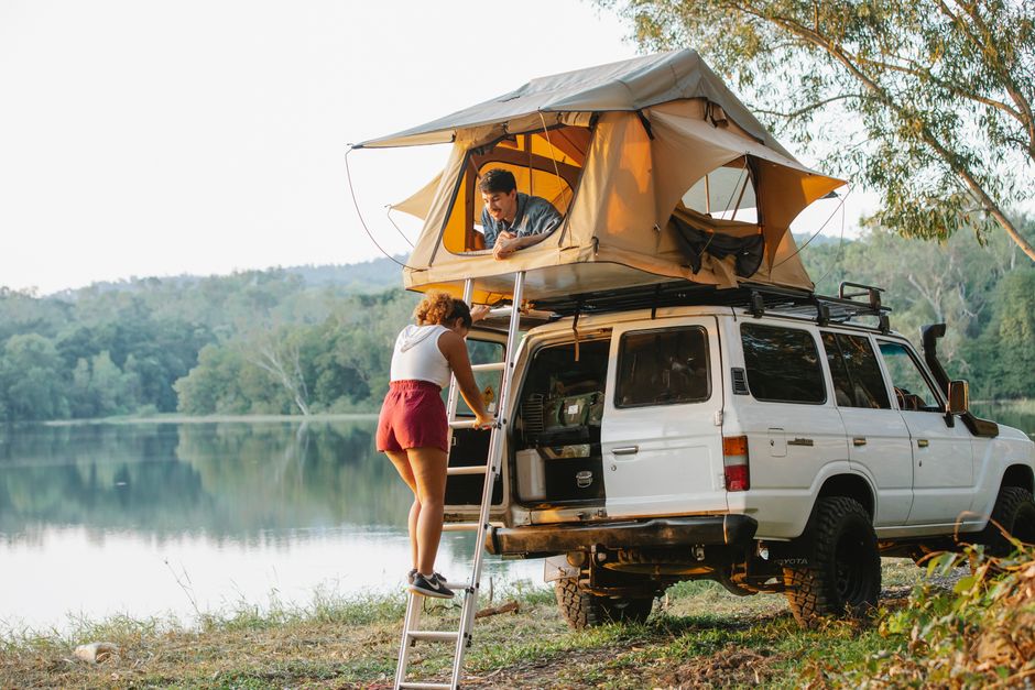 Full body of anonymous young lady ascending ladder towards positive boyfriend relaxing in camping tent laced on SUV car parked near lake during road trip