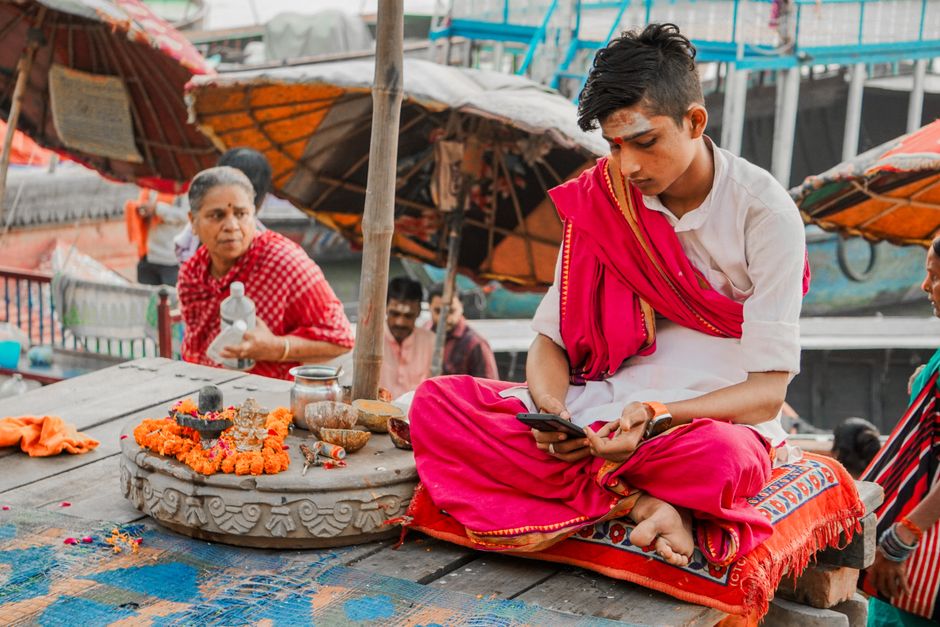 A young priest in traditional attire performs a ritual while using a phone on a riverside platform.