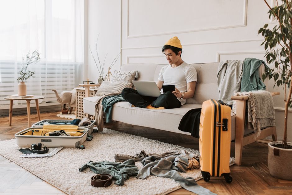 A man in a living room working on a laptop, with suitcases and clothes scattered, preparing for travel.