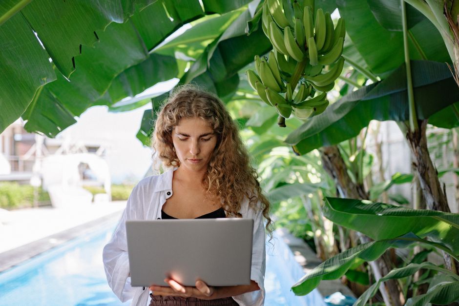 A woman with curly hair works on a laptop under lush banana trees by a poolside.