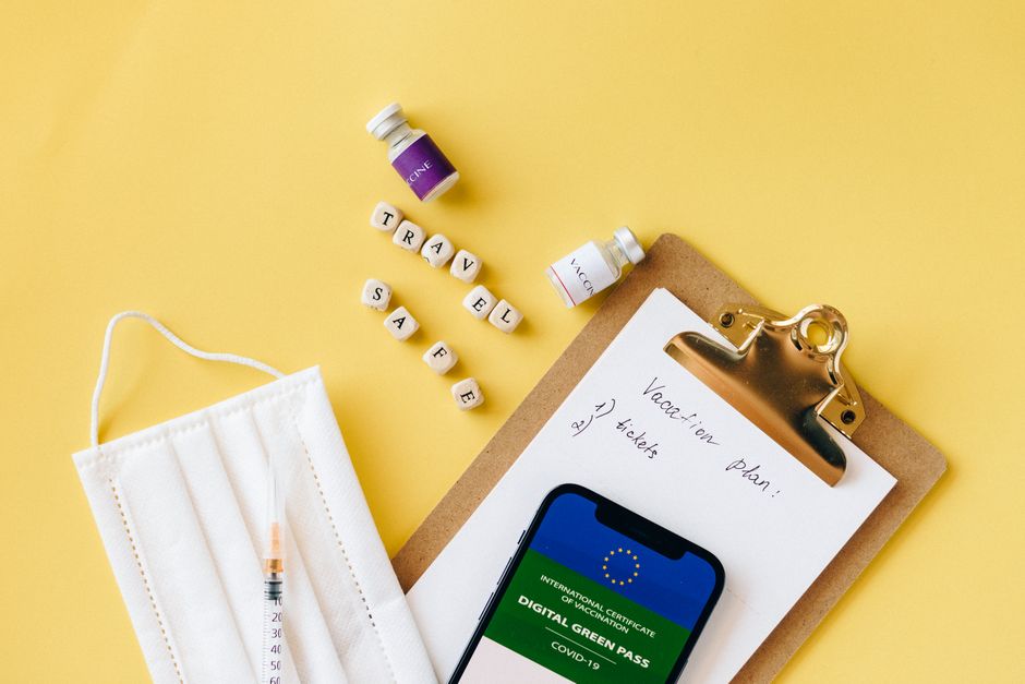 Flat lay of travel safety items: vaccine, mask, digital green pass on a vibrant yellow background.