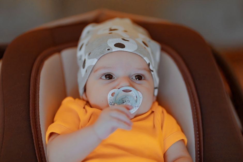 Adorable baby with beanie and pacifier in orange shirt, indoors.