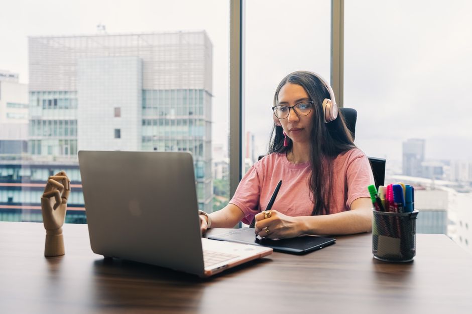 Professional woman using laptop and stylus in a modern office setting, wearing headphones.