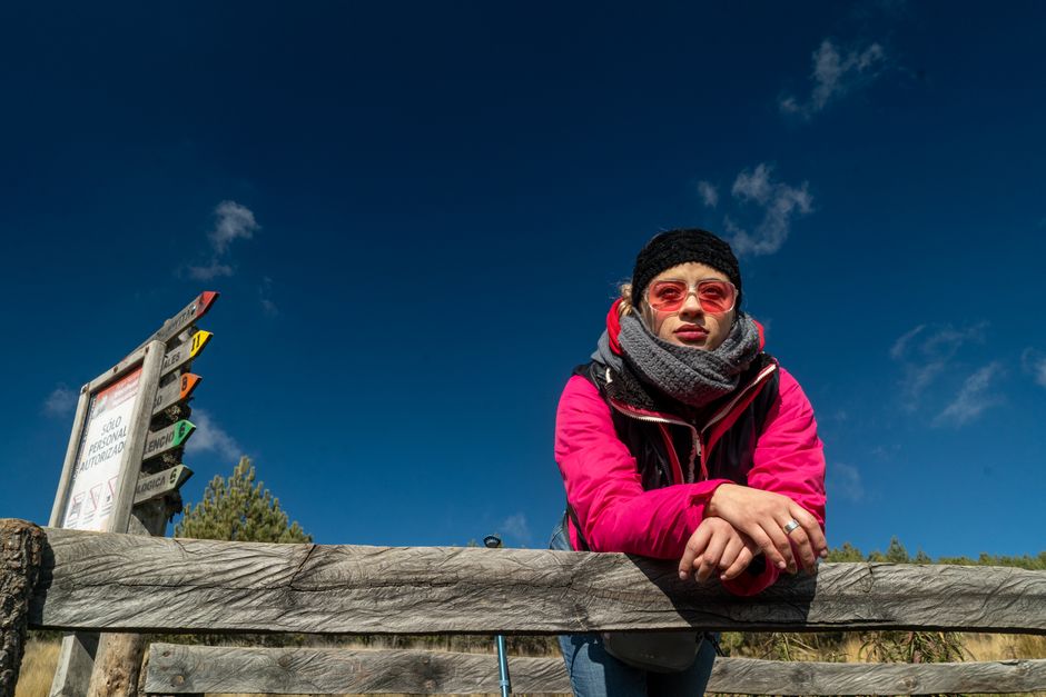 Stylish woman in winter attire standing by a rustic wooden fence on a sunny day.