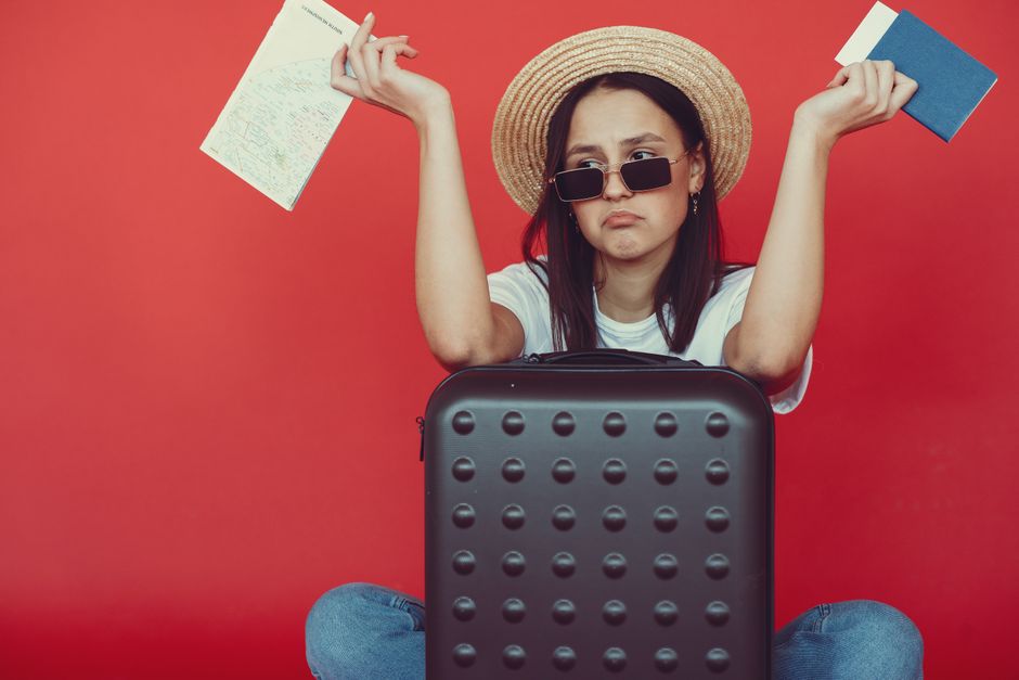 Doubters young stylish woman in wicker hat and trendy sunglasses sitting behind black suitcase and shrugging with passport and tickets on red background