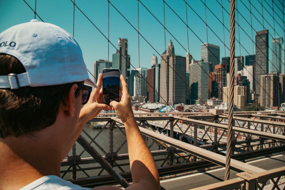 A tourist takes a photo of the New York City skyline from the iconic Brooklyn Bridge during the day.