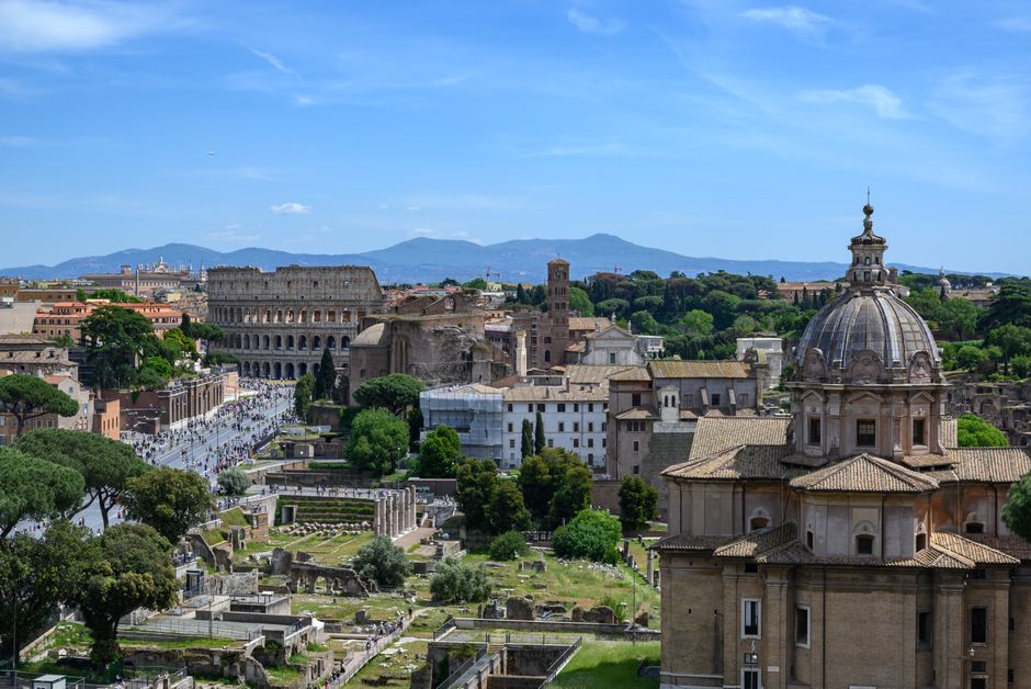 A breathtaking view of Rome with the Colosseum surrounded by historic ruins and lush greenery under a clear blue sky.