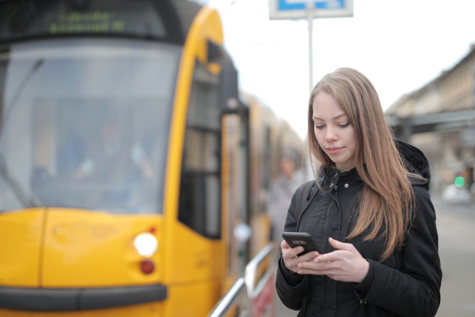A young woman using her smartphone while waiting at a city tram station during the day.