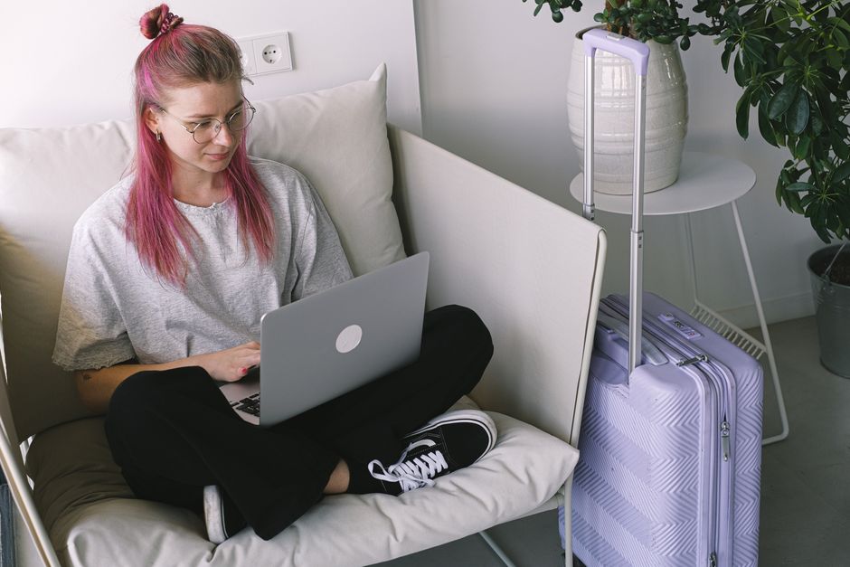 Casual young woman with pink hair works on laptop in comfortable indoor setting, travel suitcase nearby.