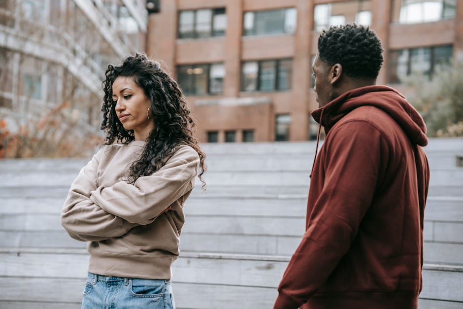 Offended ethnic girlfriend with curly hair standing with crossed arms standing near African American boyfriend while having conflict on street in park